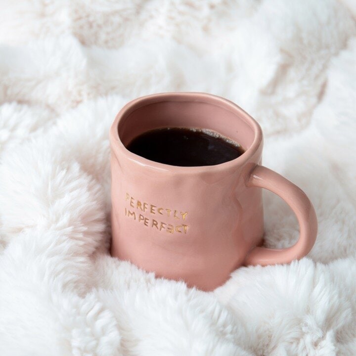 Rosa krus med teksten «Perfectly Imperfect», fylt med kaffe på et mykt hvitt teppe.