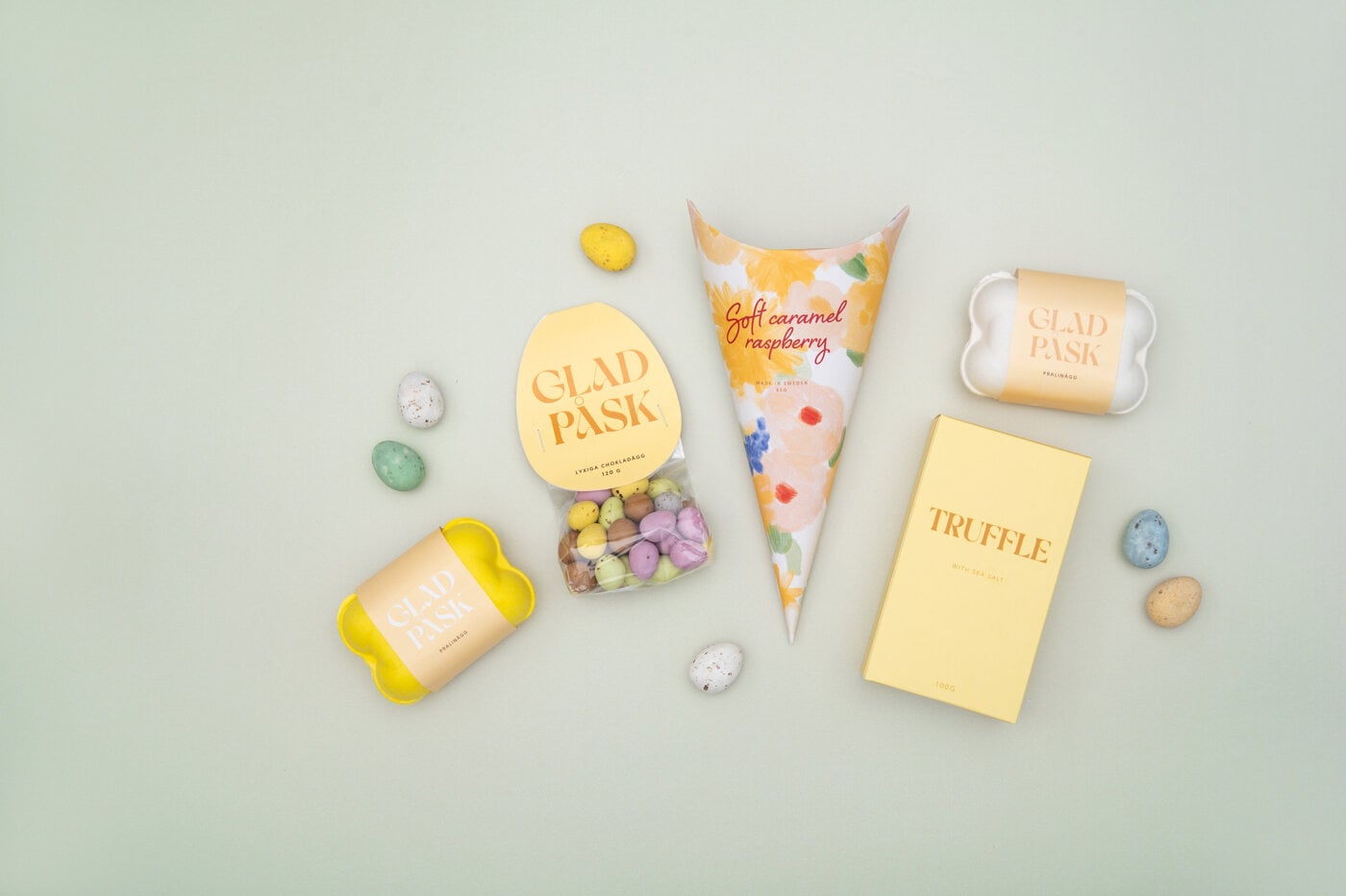 Påskegodteri i pastellfargede pakninger og sjokoladeegg mot lys bakgrunn.