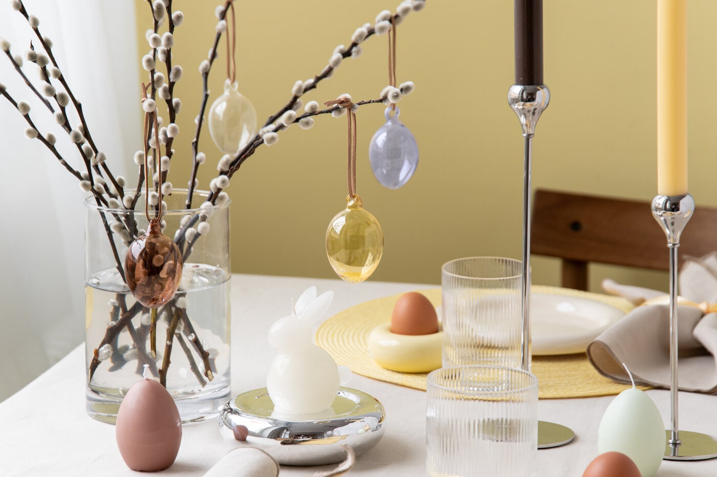 Påskedekorasjoner på bord med glassvase, seljekvister og hengende egg i myke vårfarger