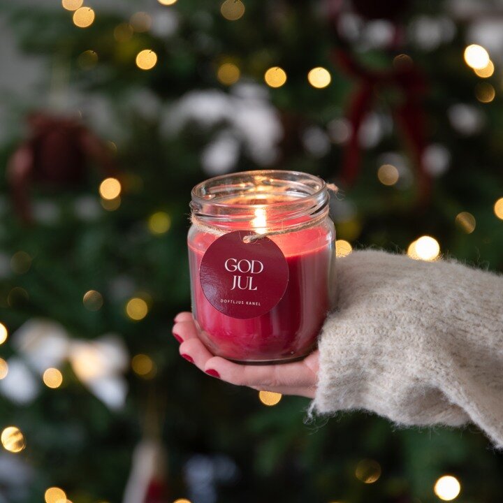 Rødt duftlys med kanel og teksten «God jul» foran et pyntet juletre.