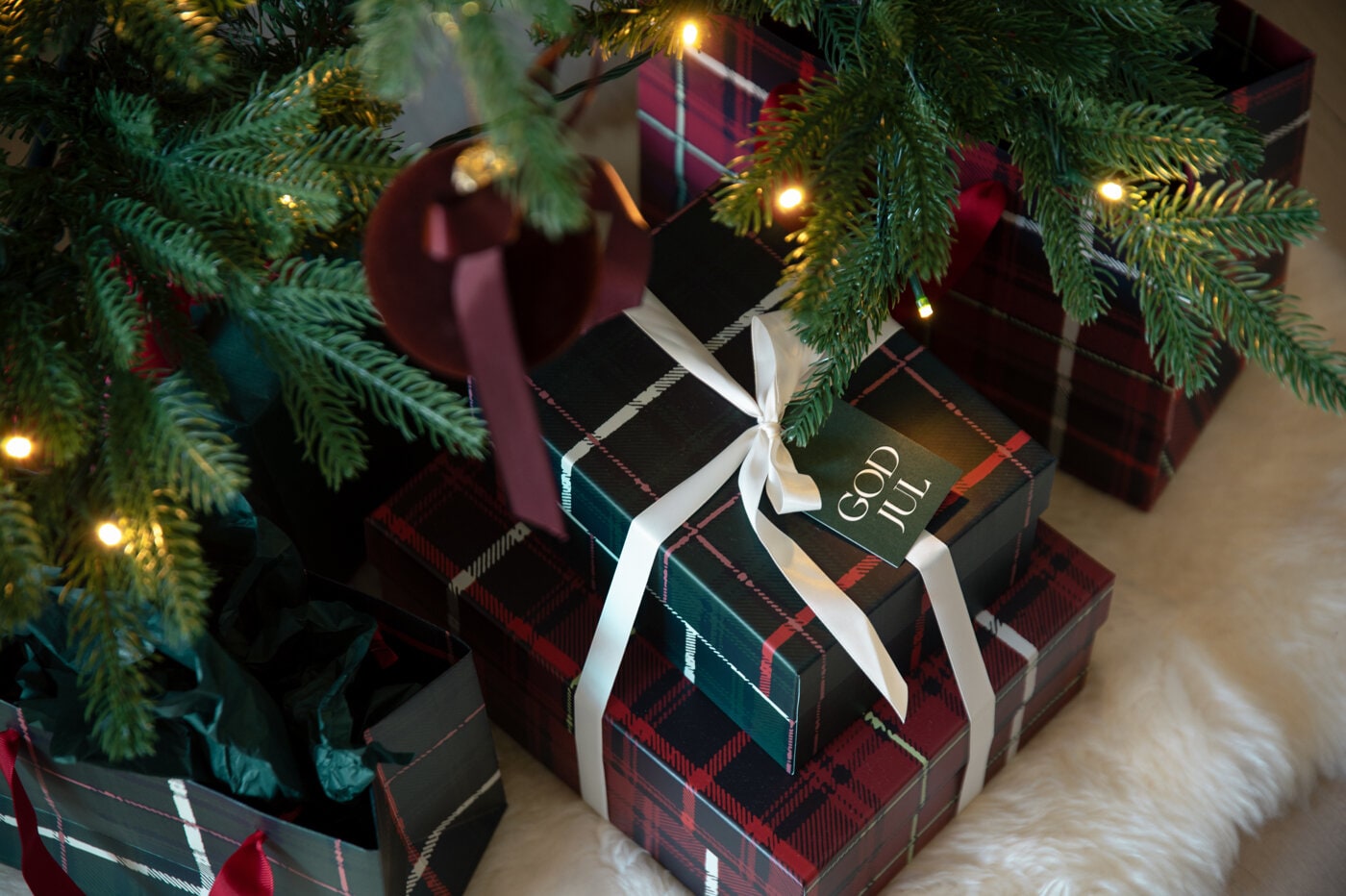 Inslagna julklappar i rutigt presentpapper med vita band under en julgran.