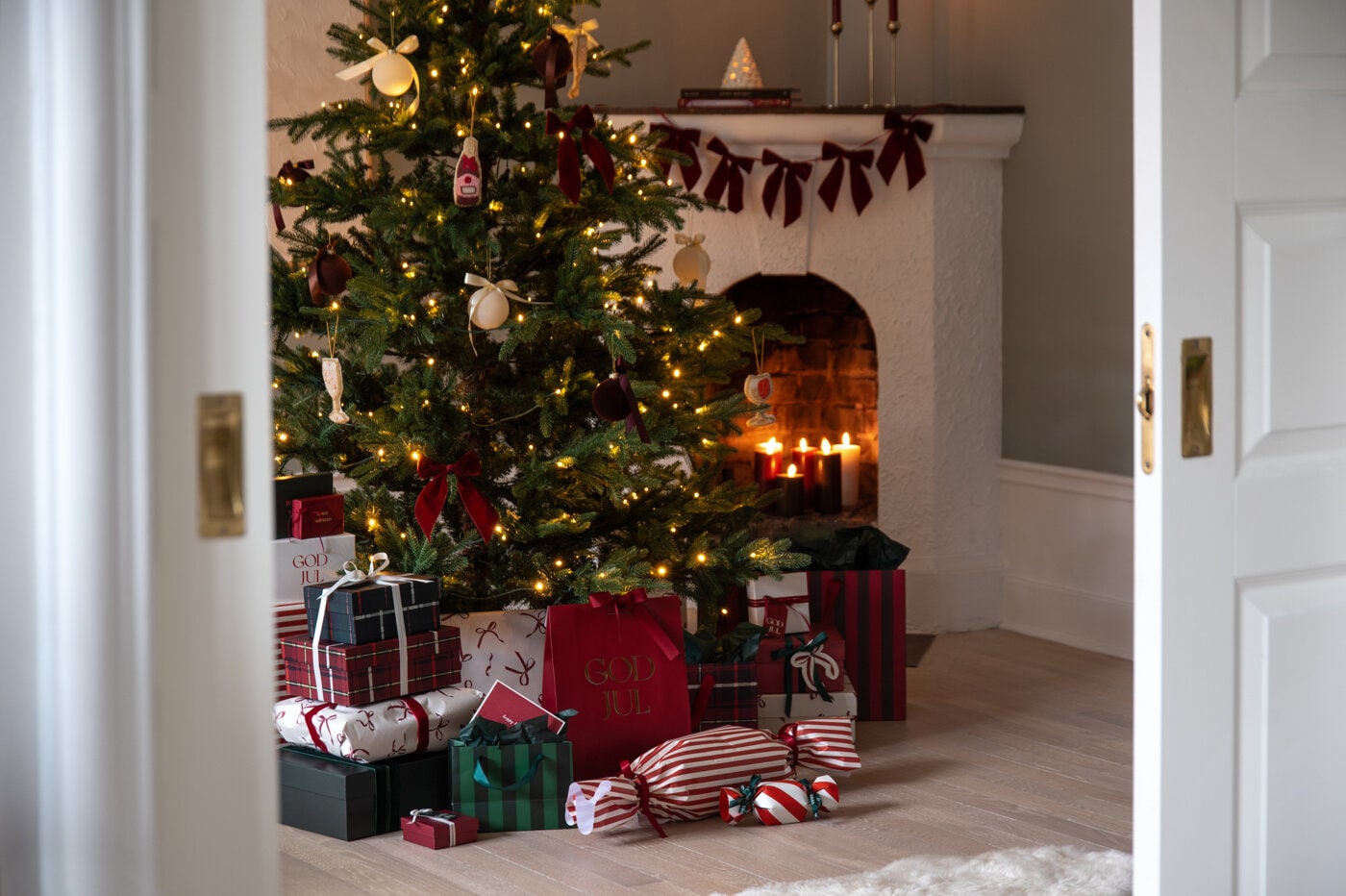 Pyntad julgran med ljusslinga och röda rosetter, omgiven av inslagna julklappar framför en öppen spis.