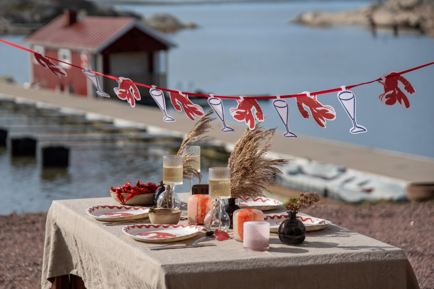 Kräftskiva dukad utomhus med kräftgirlang, tallrikar och levande ljus vid vattnet.