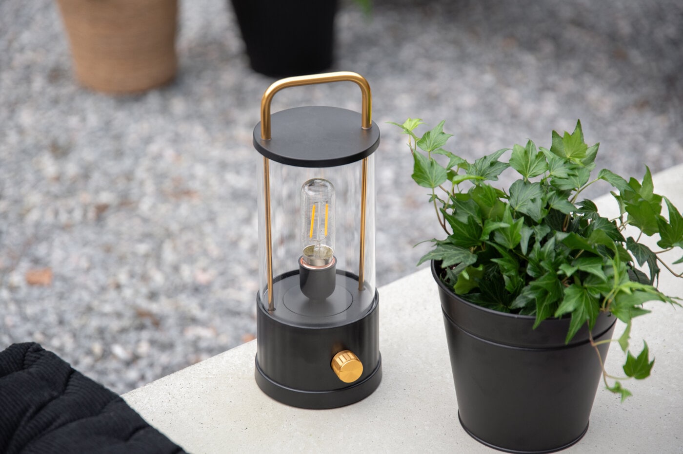 Lykt med håndtak og svart krukke med grønn plante i utemiljø.