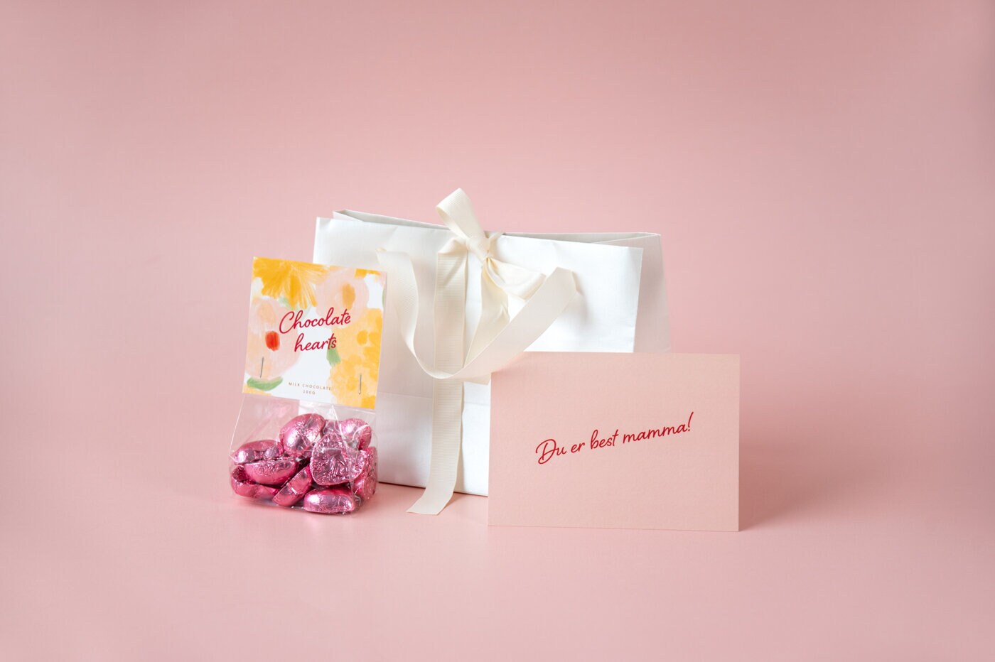 Gavesett med hvit gavepose, sjokoladehjerter og kort på rosa bakgrunn.