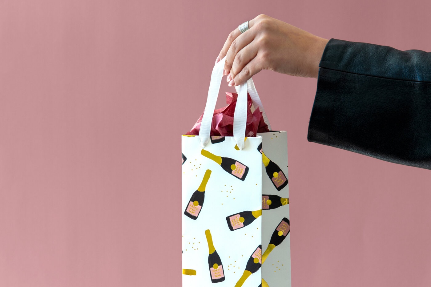 Hand som håller presentpåse med champagneflaskmotiv mot rosa bakgrund.