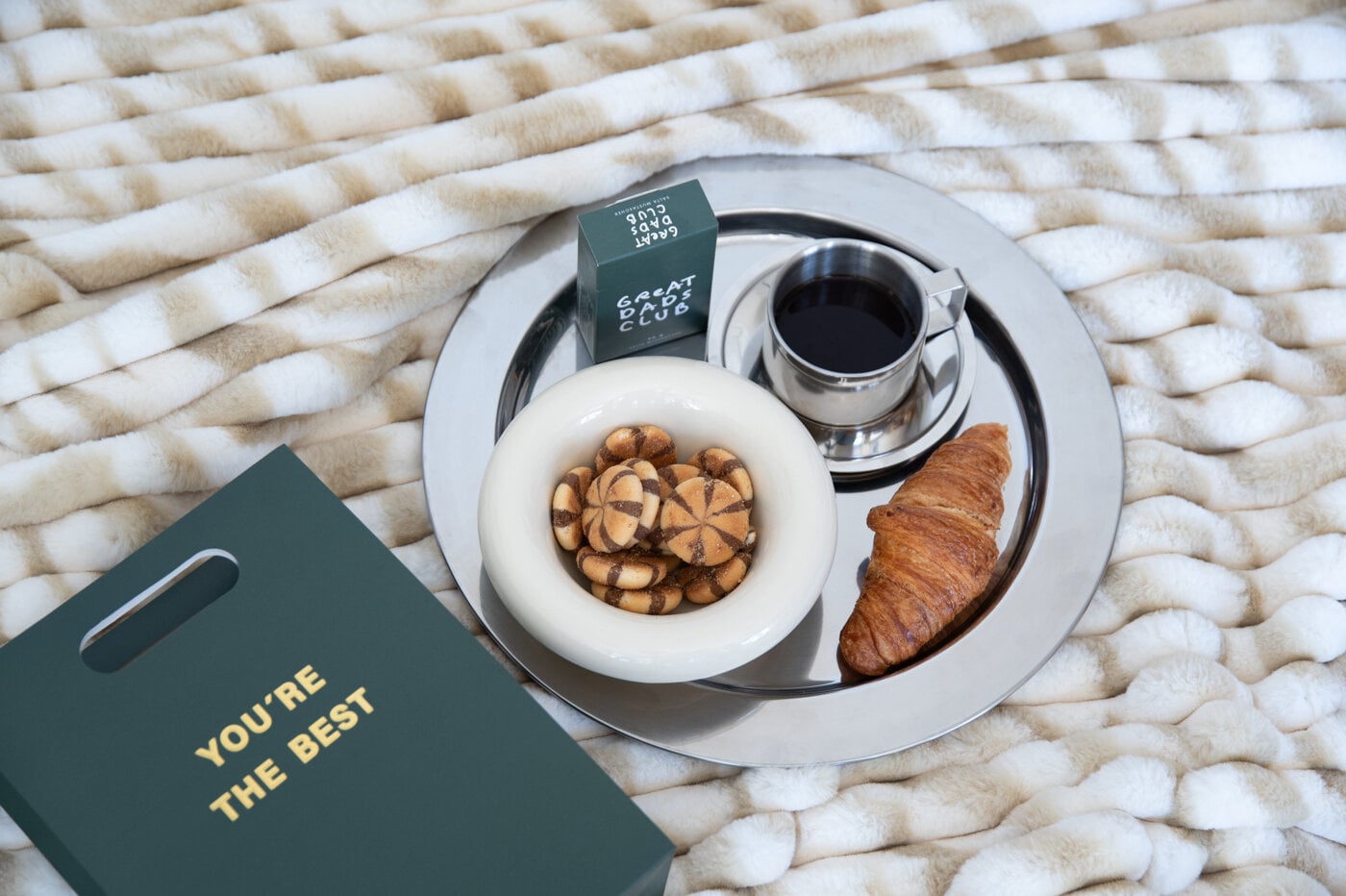 Brett med kaffe, croissant, småkaker og en gavepose på et mykt teppe.